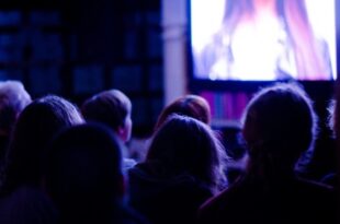 A darkened room in which we see the back of half a dozen people’s heads watching a TV screen.