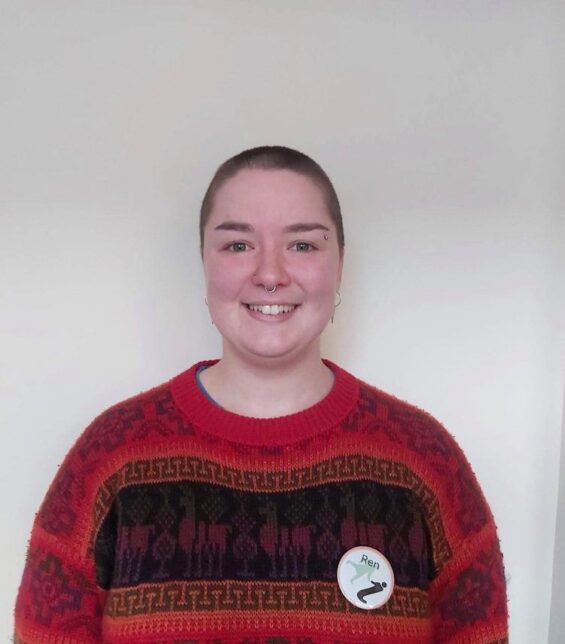 Ren stands against a white background. Ren has a cropped hairstyle and wears a red knitted wool jumper with patterns in black. Ren wears a large badge with 'Ren' written on it.