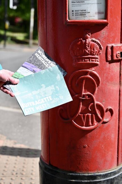 Close up of a red postbox.  A hand is the frame, holding two foldout Women's Heritage Maps