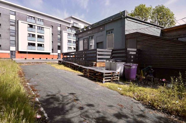 Colour photo taken on a sunny day. There is a tarmac road leading up to a grey brick modern block of flats.