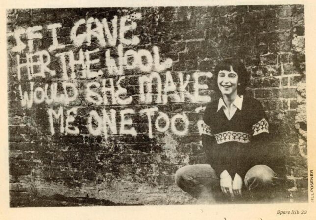 Monochrome photograph of a woman wearing the Spare Rib 'woman's woolly' knitted jumper, squatting beside a wall sprayed with the words 'If I gave her the wool would she make me one too?'