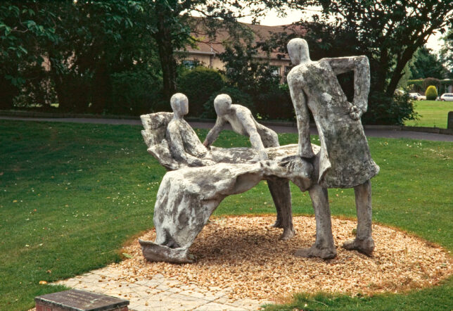 a metal sculpture by Edith Simon showing three figures, two of whom are standing, the third is lying in a bed which appears to levitate