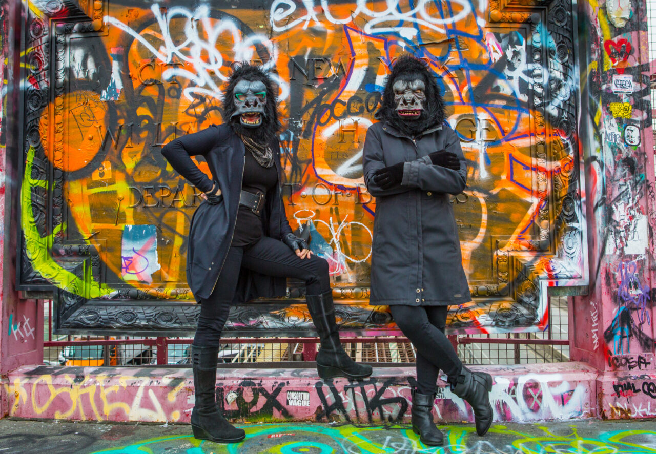 Two figures are photographed standing against a wall which is daubed with colourful graffiti. The figures are dressed in dark clothing and each is wearing a fancy dress style gorilla mask