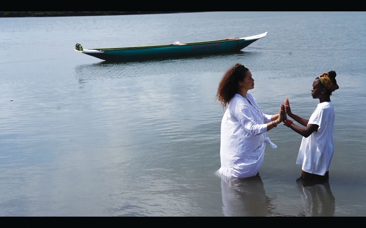 Alberta Whittle, Still from Holding the Line, 2020. Two women of colour stand in deep water, facing each other with their hands pressed together. A small low boat floats in the water behind them.