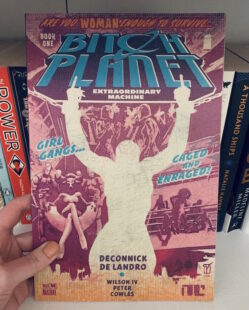 Image shows cover of Bitch Planet. A silhouette of a woman’s body is visible and she is giving two fingers. To the right hand side of the silhouette a woman is sitting on a chair with bodyguard type people behind her. To the right hand side women are standing and sitting next to a railing. Picture is taken against the backdrop of a bookcase.