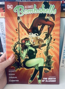 Image shows Bombshells cover with Poison Ivy sat on a swing covered in vines, wearing a green bodice. Harley Quinn hangs above her wearing a harlequin outfit. Picture is taken against the backdrop of a bookcase.