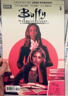 Image shows cover of Buffy the Vampire slayer issue 2. Buffy is sat at a desk with books looking at the reader and Drusilla stands behind her in a pink circle.