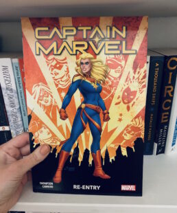 Image shows cover of Captain Marvel standing in a power pose wearing her uniform. Background lights show past depictions of Captain Marvel shining through. Picture is taken against the backdrop of a bookcase.