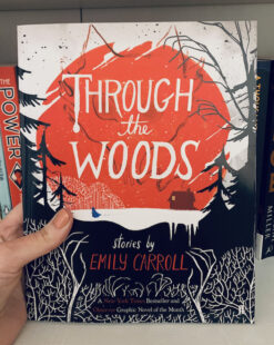 Image shows cover of Through the Woods and depicts a black and white forest with a red background that has ‘Through the Woods’ and a house within it. Picture is taken against the backdrop of a bookcase.