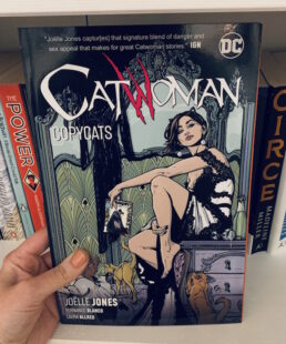 Image shows cover of Catwoman sitting on a dresser holding a Batman comic. She wears an evening gown and her costume hangs over a mirror in the back. She is surrounded by cats. Picture is taken against the backdrop of a bookcase.
