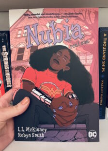 Image shows cover of Nubia sitting looking at the reader. She is wearing a Wonder Woman t-shirt and bracelets of submission. Picture is taken against the backdrop of a bookcase.