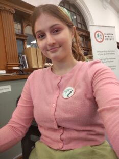 Myriam, a volunteer, wearing a GWL badge. She is setting at a desk and a GWL banner is visible behind her.