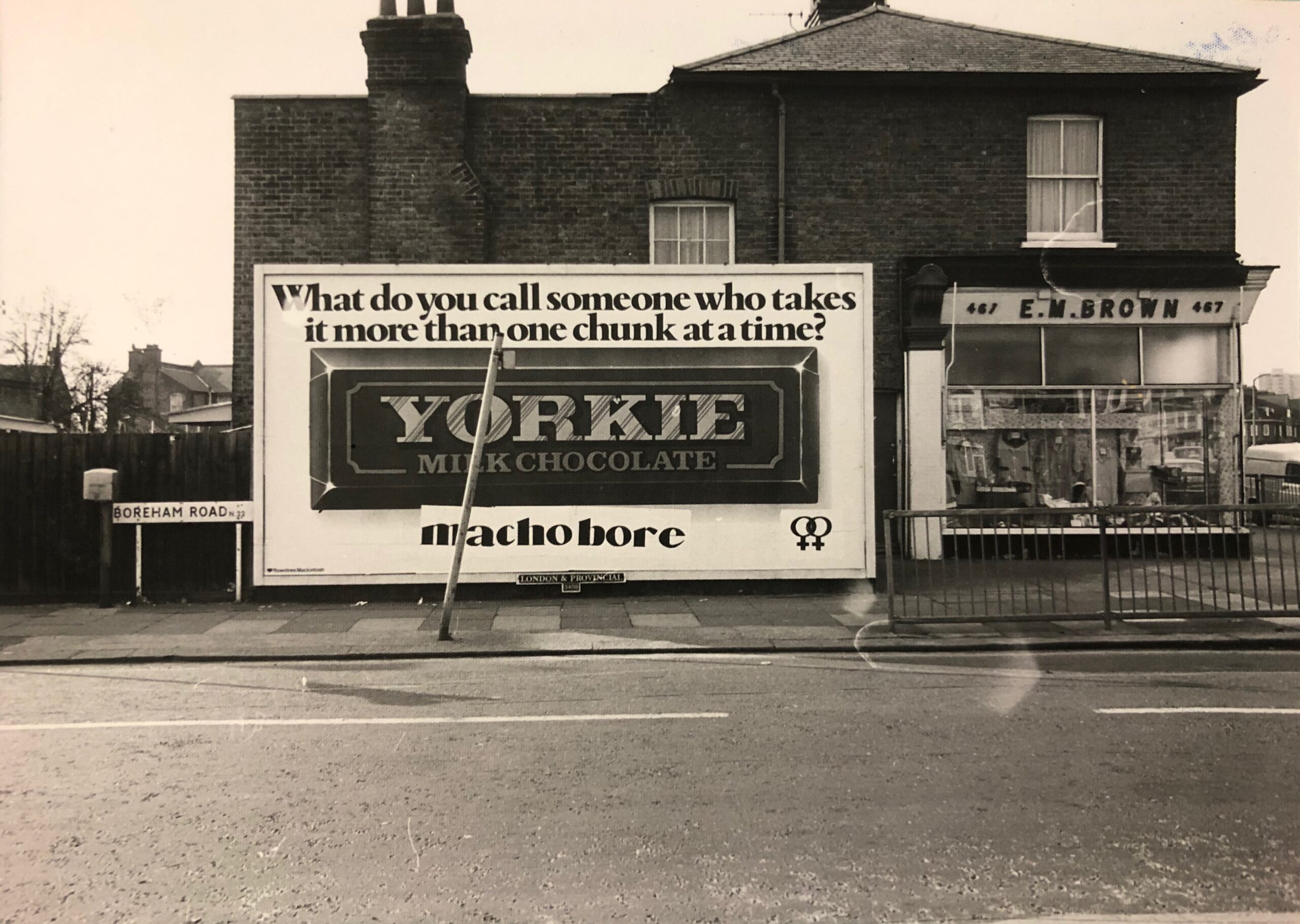 Billboard advert for Yorkie chocolate, featuring a Yorkie bar in the centre and, 'What do you call someone who takes it more than one chunk at a time?' above. Below the bar, someone has wheat-pasted 'macho bore' and the lesbian symbol. 