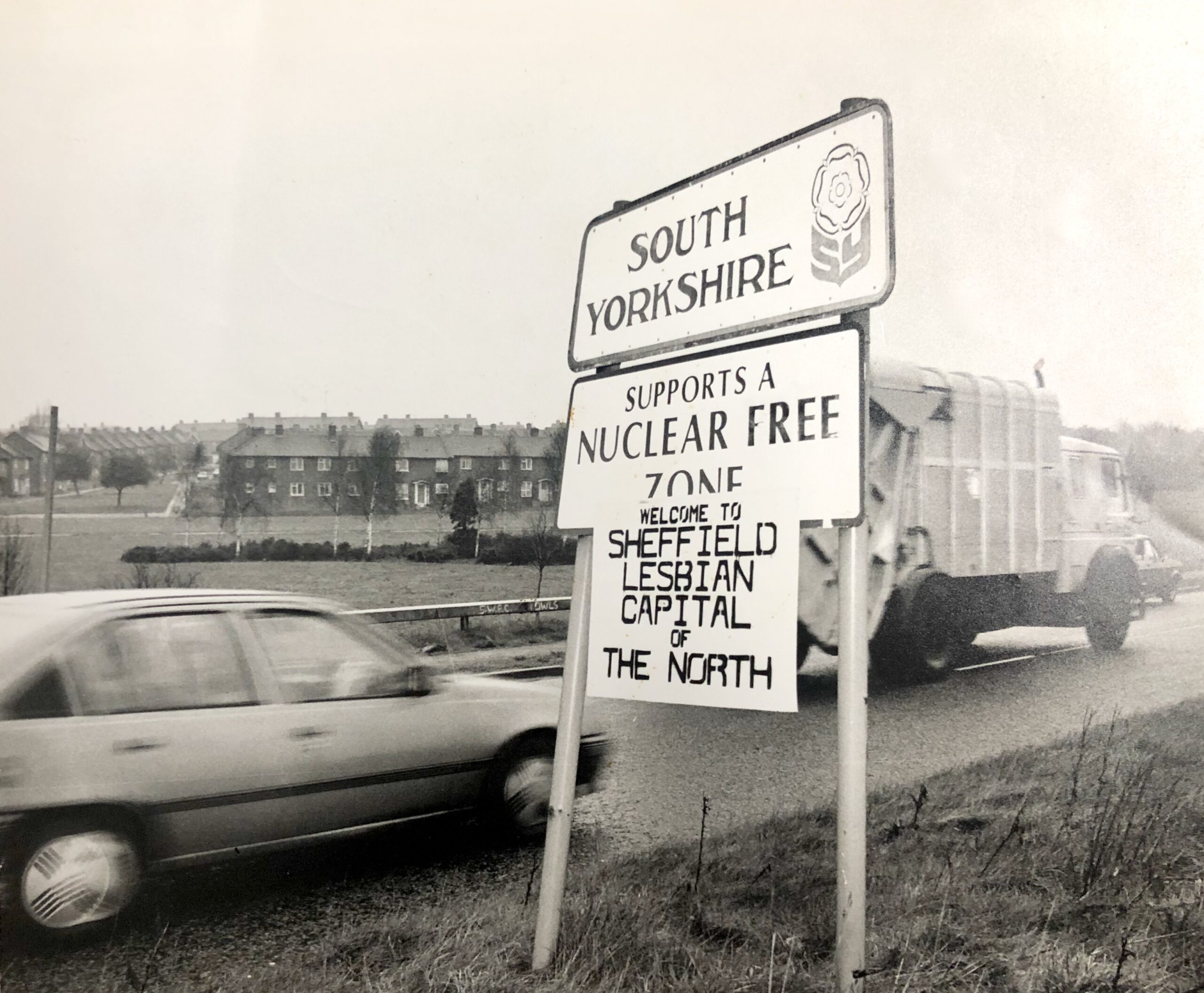 Boundary sign for South Yorkshire, to which someone has affixed a placard that reads 'Welcome to Sheffield lesbian capital of the north'. Houses are visible in the background, while a car drives past in the foreground. 