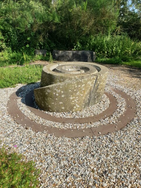 A photograph of the grey stone and steel circular water fountain in the center of a spiral surrounded by little grey stones on the ground. There is flora and fauna in the background.