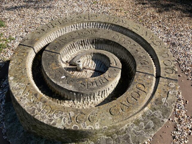 Close up photograph of the grey circular steel and stone water fountain with the words "Women's Peace Camp 1981 to 2000" and "You can't kill the Spirit" engraved into it. 