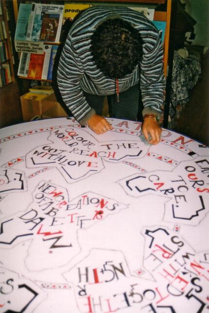 A woman leans over a large round table, painting colourful slogans including 'Colour the World, Reach Out With Love'