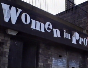 Women in Profile sign, white text on black background, outside a brick building.