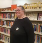 A colour photo of Helen. Helen is standing in front of a shelf filled with kids books at the library. Helen is wearing a black dress with a long black beaded necklace. Helen is smiling, she has on glasses and her hair is tied back.