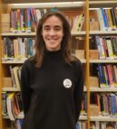 A photo of Kirstin. Kirstin wears a black roll neck top and wears a round badge with their name on it. Kirstin is smiling and standing in front of a bookshelf filled with books