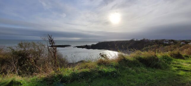 A photo taken of Catterline beach near the Watchrie. The cliff edge is full of grassy greenery and curves around to the right. The sea looks still and grey. The sky has lots of grey clouds but the sun is poking through. 