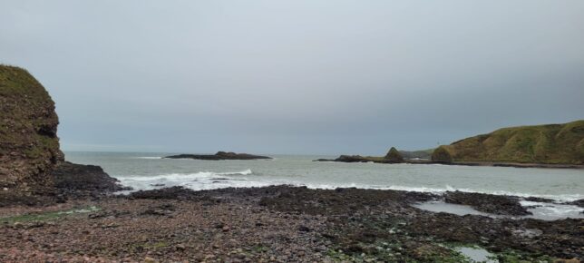 Photo of Catterline beach. It is a stone beach rather than sand and the grassy cliffs are on the right. The water is greyish blue with white waves and the sky is a pale grey.