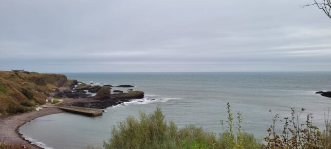 A photo of Catterline beach where you can see the pier. It is a stone beach rather than sand and the sea is grey. The sky is also grey and the Watch Tower can be seen on top of a grassy cliff to the left. 