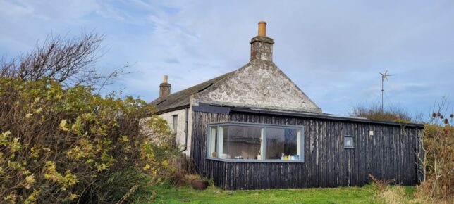 Photo of a small house made from grey stone and wood panels painted black. 