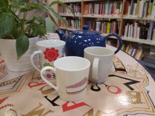 Photo of a big blue tea pot and three mugs sitting on a colourful table.