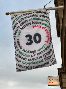 GWL's 30th anniversary flag flying from the pole outside the library. The flag features a large "30" in the centre, spiralling outwards, in red text, are the words "years of changing" followed by a number of other words in green and black text including "minds", "perspectives", "Museums", "Libraries" and "Glasgow"