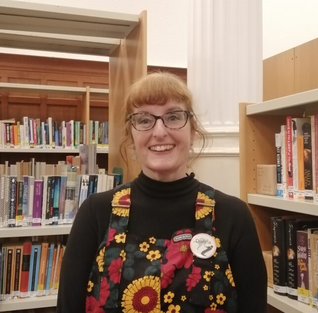 A colour photo of Donna Moore. Donna is wearing a colourful pair of dungarees emblazoned with yellow and red flowers. Donna's hair is red and is tied back. Donna wears tortoiseshell glasses and has a big grin on her face.