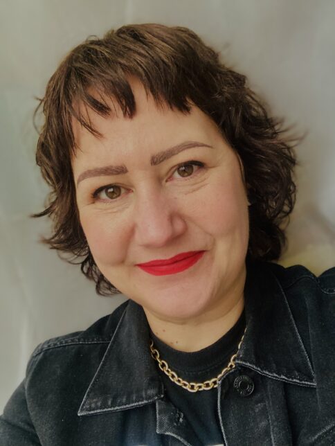 A colour photo of Rachel Thain-Gray. Rachel has short, dark hair and wears red lipstick, she smiling at the camera. Rachel wears a cold curb chain, a black t-shirt and black denim jacket