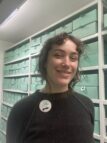 A Colour photograph of Mae Moss. Mae is pictured standing in the archive store, Mae is standing in front of green archive boxes on shelves. Mae wears a black sweater with a Glasgow Women's Library badge with her name on. Mae is smiling and looking at the camera