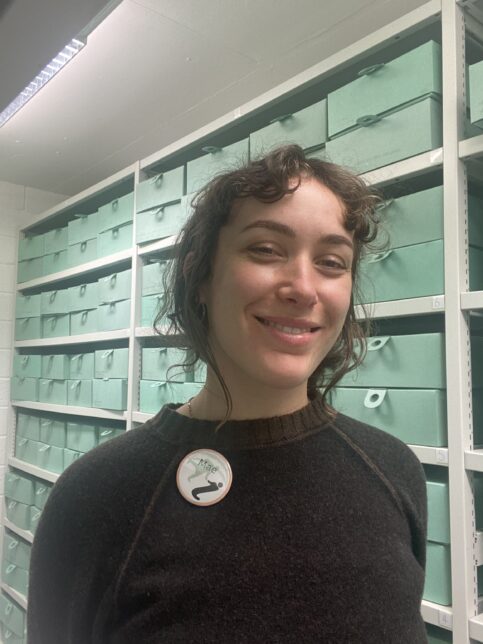 A Colour photograph of Mae Moss. Mae is pictured standing in the archive store, Mae is standing in front of green archive boxes on shelves. Mae wears a black sweater with a Glasgow Women's Library badge with her name on. Mae is smiling and looking at the camera