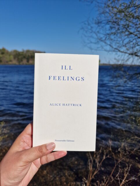 A photo showing Naomi's hand holding the book 'Ill Feelings' by Alice Hattrick, published by Fitzcarroldo Editions. The book has a white cover with blue text on the front. Naomi is holding the book up in front of an expansive loch under blue skies.