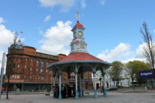 A group is standing under the Bridgeton Umbrella, an open shelter in the main square. The shelter is open on all side and has a red roof and blue structure. 