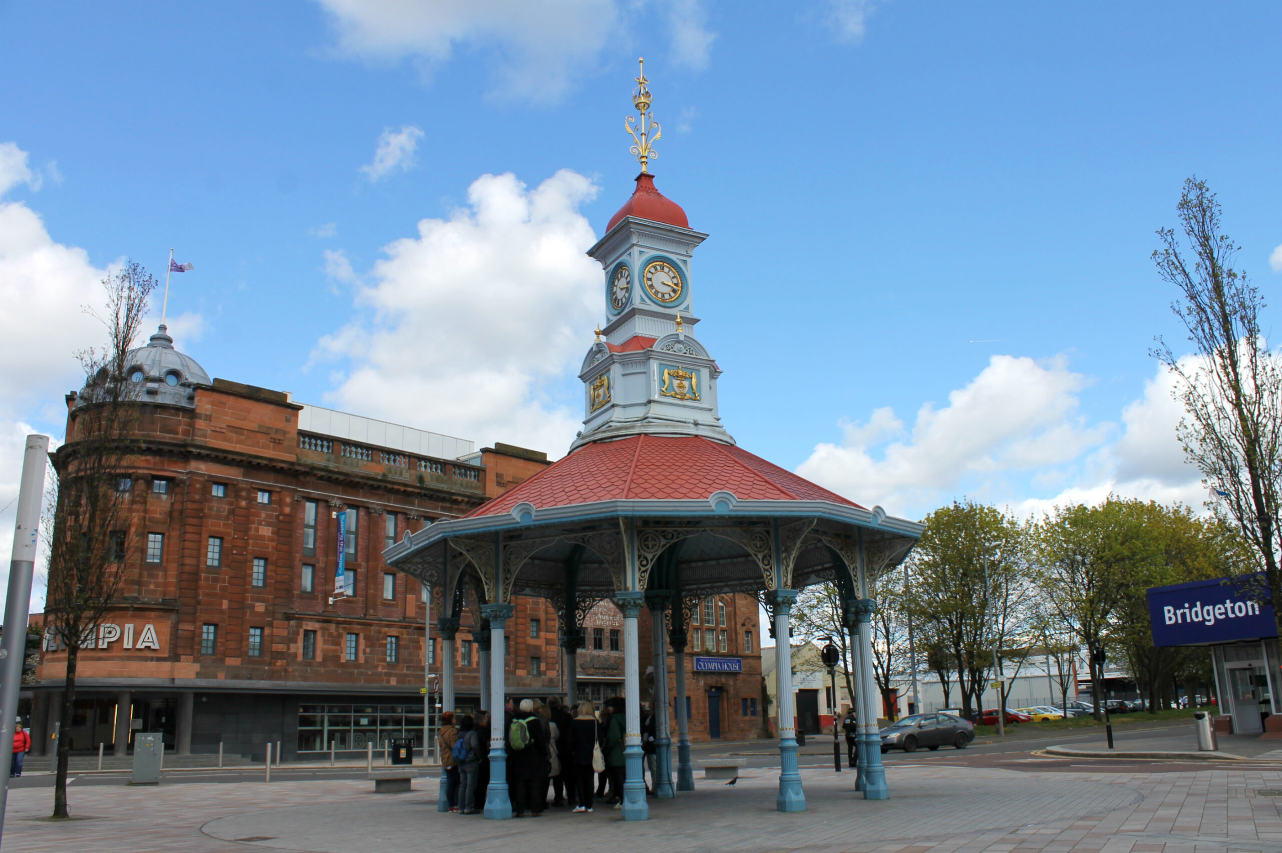 A group is standing under the Bridgeton Umbrella, an open shelter in the main square. The shelter is open on all side and has a red roof and blue structure.