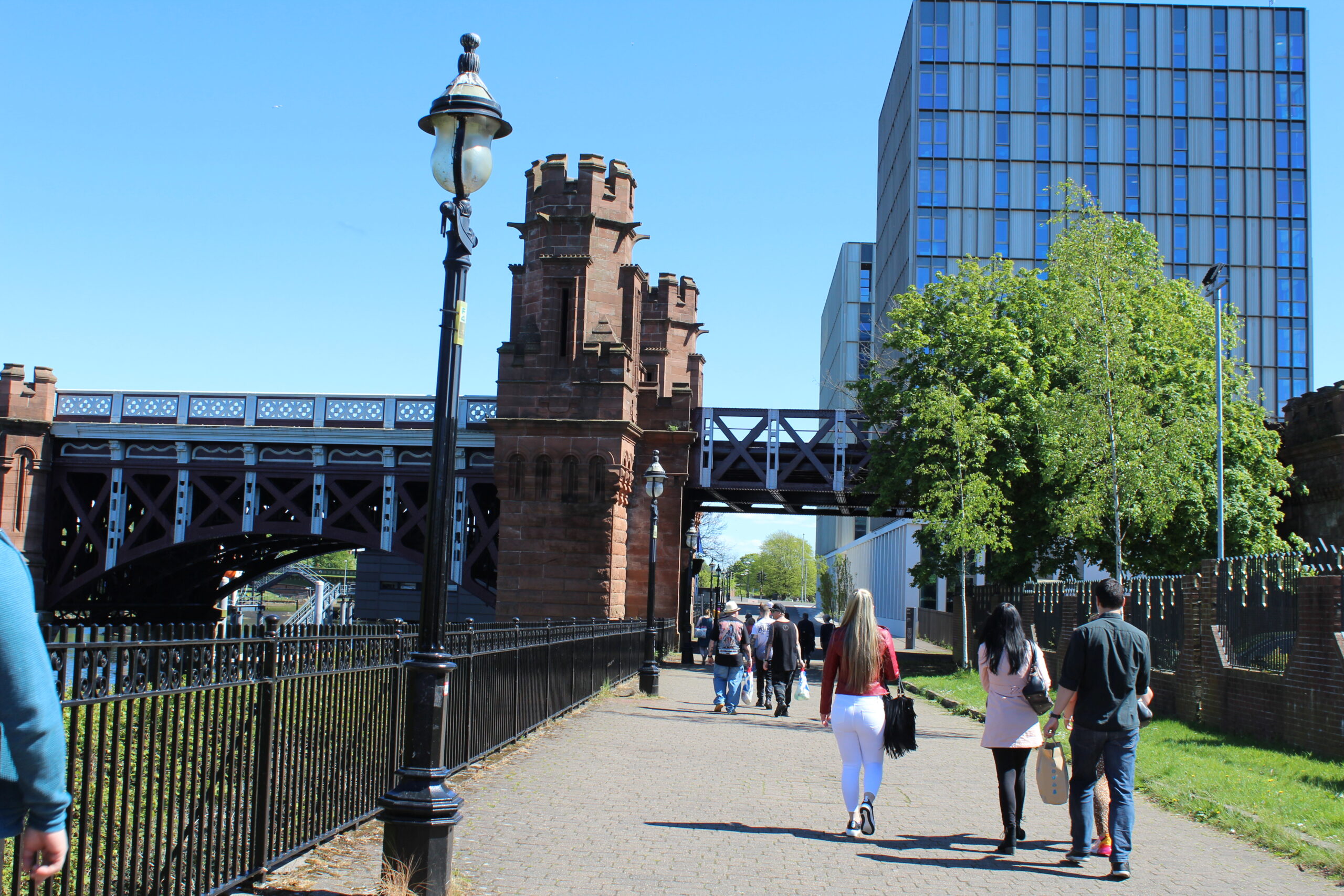 It’s a sunny day with a cloudless blue sky. People are strolling along a walkway by the side of the river Clyde. There is a bridge and tower block in the background.