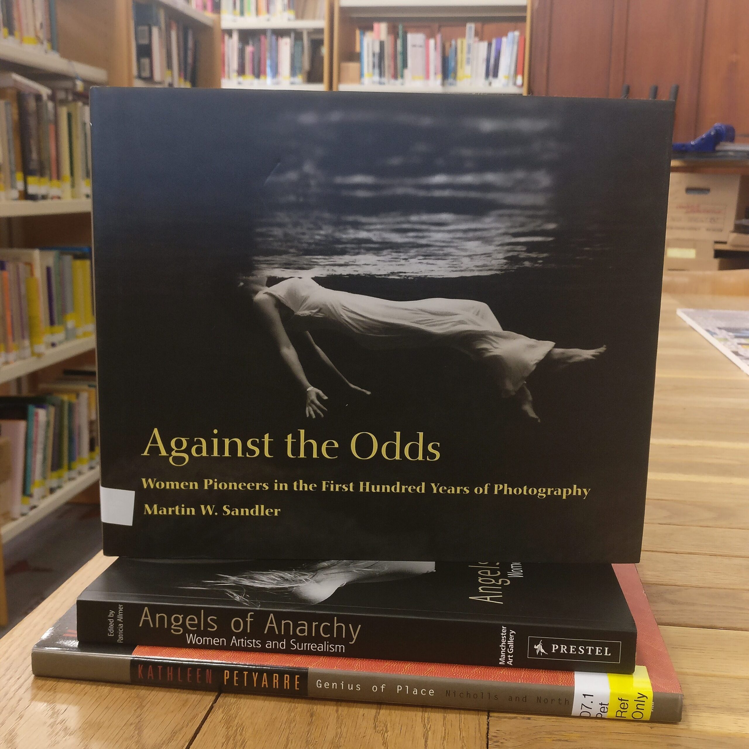 Depicting the front cover of 'Against the Odds', a black and white image of a woman in a white dress underwater, her face just breaking the surface, the title of the book below her figure. 