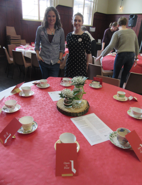 Two women stand proudly and smiling as they face the camera. They are behind a large round table with a red table cloth that is set for afternoon tea. There are red postcards propped up on stands in front of each place around the table and the postcards state Open the Door. Behind them women are busily setting up more tables.