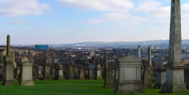 It’s a clear winter day. This is the view of the city of Glasgow from the top of a hill. In the foreground there are large tomb stones and obelisks. In the middle and back ground there are rows of houses, large building and tower blocks.