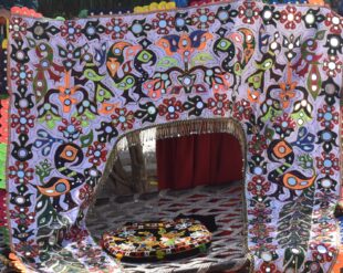 a brighty coloured hanging fabric depicts abstract bird and flower forms in oranges, blues, reds and blacks against a white background. Behind the fabric there is a room with more fabric art works on cushions and the floor. 