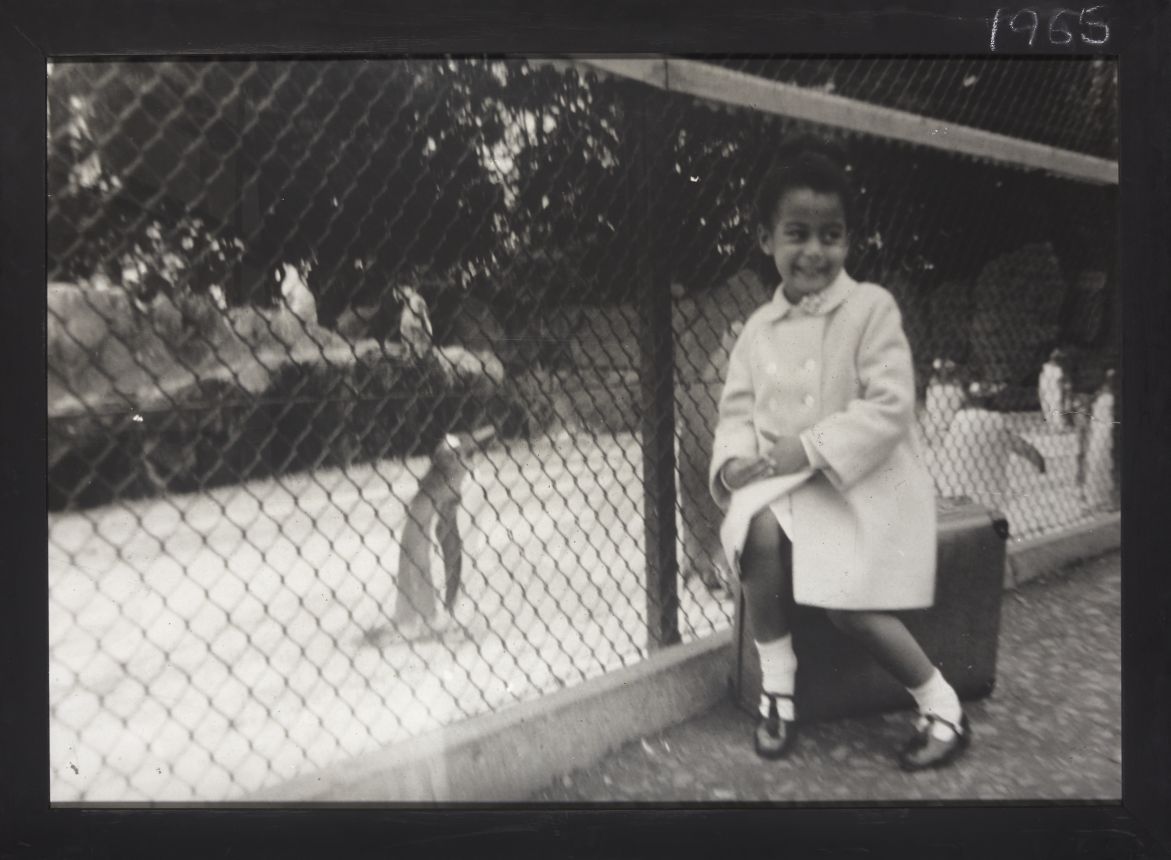 The image is a back and white photograph of Maud Sulter as a young child at Edinburgh zoo, sitting on a suitcase in front of the penguin enclosure. Sulter is wearing a coat which buttons down the front, and ankle socks with t-bar shoes. The image is presented in a black wooden frame with chalk writing in the top right hand corner with the date ‘1965’.