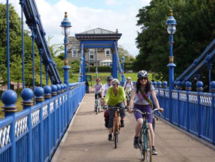 Five women are cycling leisurely towards us over a pedestrian bridge. It’s a summer day and the sky is blue.