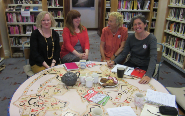Four women sit around a brightly painted table surrounded by shelves of books. They are smiling and have notebooks, tea cups and biscuits in front of them.