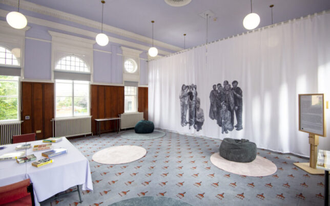 The community room at the library with benabags on the floor and a large white curtain in the background, the curtain has large illustrations by the artist Oliva Plender on