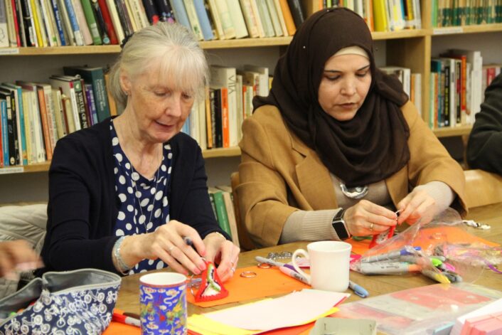 Two women sit next to each other sitting at a table making key rings with knitted pattern