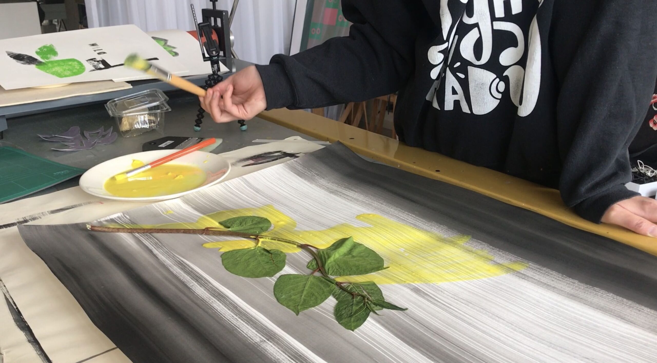 The bottom torso of a person can be seen at a table, they hold a paintbrush and our painting over some leaves onto paper