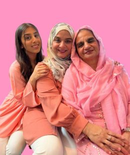 Three women hold each others shoulders, smiling. They wear pink and sit against a pink background.