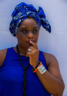 Photograph of a woman with a finger over her mouth. She wears a blue top, a beaded necklace, bangles and a blue headscarf
